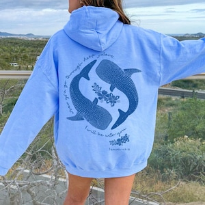 K&ouml;nnte beinhalten: Ein hellblauer Kapuzenpullover mit einer dunkelblauen Grafik auf der R&uuml;ckseite. Das Design zeigt zwei Walhaie in kreisf&ouml;rmiger Anordnung mit floralen Akzenten und dem Text: When you go through deep waters I will be with you. Isaiah 43:2.