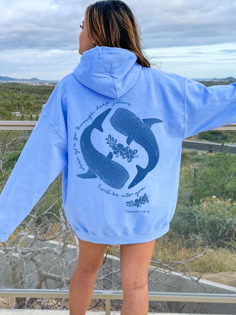 K&ouml;nnte beinhalten: Ein hellblauer Kapuzenpullover mit einer dunkelblauen Grafik auf der R&uuml;ckseite. Das Design zeigt zwei Walhaie in kreisf&ouml;rmiger Anordnung mit floralen Akzenten und dem Text: When you go through deep waters I will be with you. Isaiah 43:2.