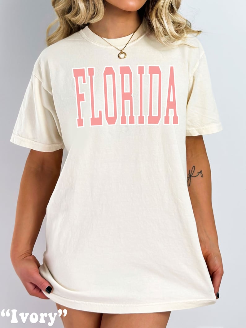 Op de afbeelding: Een wit T-shirt met een roze "FLORIDA" print op de voorkant. Het shirt wordt in de afbeelding beschreven als "Ivoor".