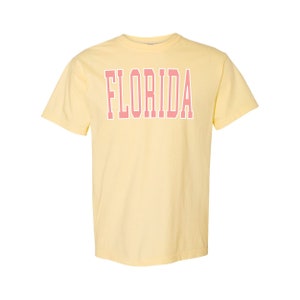 Op de afbeelding: Een geel T-shirt met korte mouwen met het woord "FLORIDA" in roze op de voorkant.