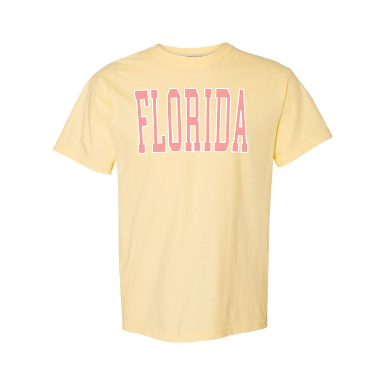 Op de afbeelding: Een geel T-shirt met korte mouwen met het woord "FLORIDA" in roze op de voorkant.