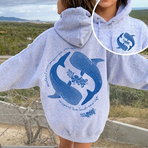 K&ouml;nnte beinhalten: Ein graumelierter Kapuzenpullover mit einer gro&szlig;en blauen Grafik auf der R&uuml;ckseite, die zwei Walhaie in einem Kreis mit Hibiskusbl&uuml;ten und dem Text "protect our precious oceans respect the local sea life" zeigt. Eine kleinere passende Grafik befindet sich auf der Vorderseite.