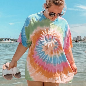 May include: A tie-dye t-shirt with a large sun graphic. The shirt features a spiral design in shades of blue, green, pink, and orange. The sun graphic is white with a face in the center. The person is holding white sandals with silver studs.