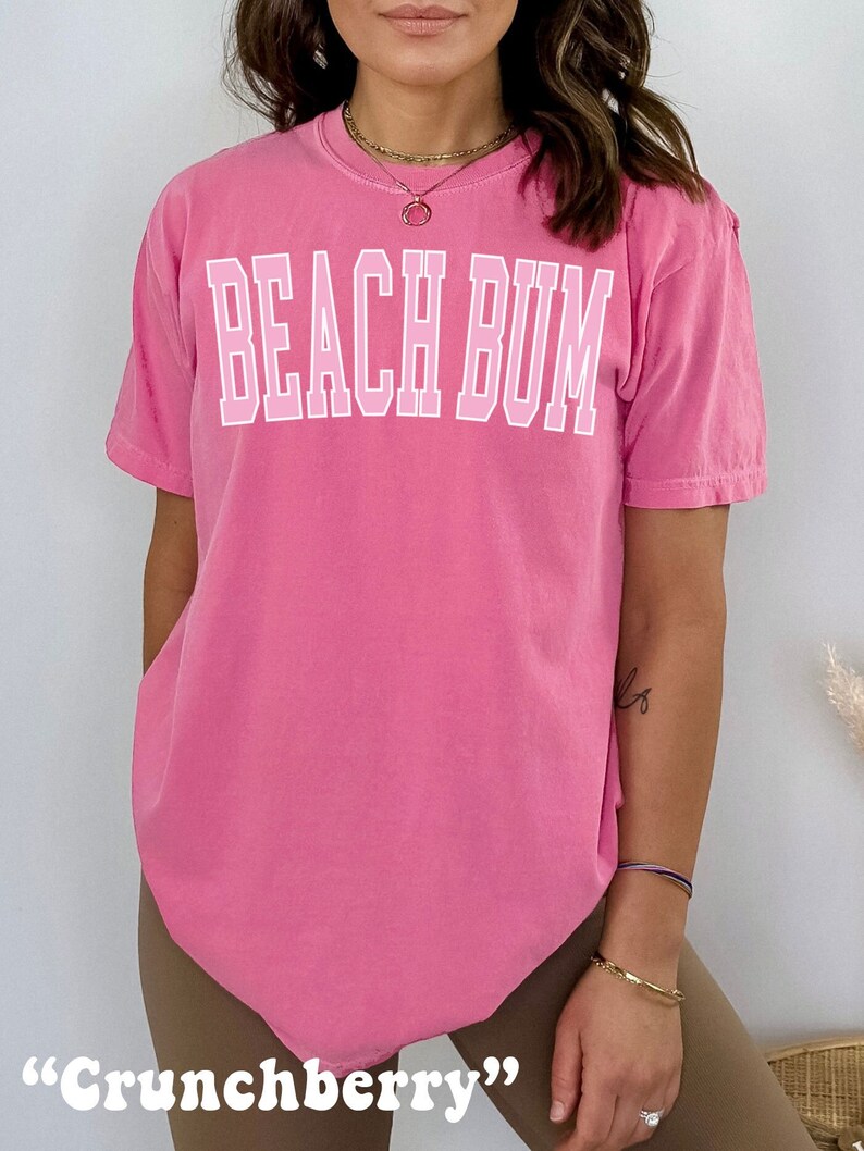May include: A pink t-shirt with the text "BEACH BUM" in white outlined letters. The shirt is a relaxed fit and has a crew neck.