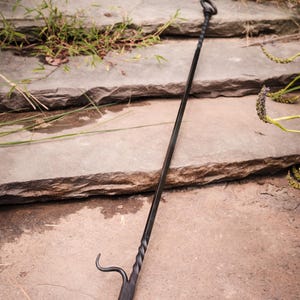 May include: A black wrought iron fire poker with a twisted shaft and a pointed end with a hook. The top has a loop for hanging. The poker is resting on stone steps with grass and plants.