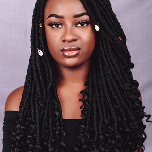 Könnte beinhalten: Eine Frau mit langen, schwarzen Kunst-Locs, verziert mit kleinen goldenen Perlen und weißen Muschelakzenten. Das Haar fällt herab, mit lockeren, lockigen Enden. Sie trägt ein schwarzes schulterfreies Oberteil und hat ein neutrales Make-up.