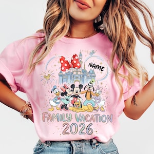 May include: A light pink t-shirt featuring a colorful graphic of cartoon characters including Mickey Mouse, Minnie Mouse, Donald Duck, Daisy Duck, Goofy, and Pluto in front of a castle. The text reads "NAME", "FAMILY VACATION", and "2026".