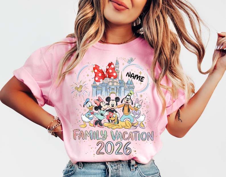 May include: A light pink t-shirt featuring a colorful graphic of cartoon characters including Mickey Mouse, Minnie Mouse, Donald Duck, Daisy Duck, Goofy, and Pluto in front of a castle. The text reads "NAME", "FAMILY VACATION", and "2026".