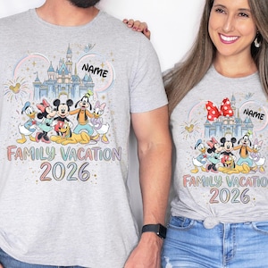 May include: Two light gray t-shirts featuring a colorful graphic of a castle, fireworks, and Disney characters including Mickey, Minnie, Donald, Daisy, Goofy, and Pluto. The text reads "NAME", "FAMILY VACATION", and "2026" in a rainbow font.