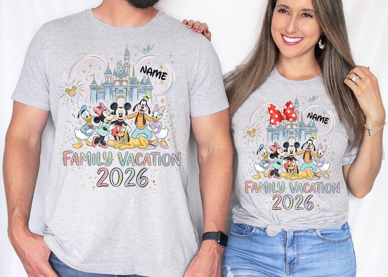 May include: Two light gray t-shirts featuring a colorful graphic of a castle, fireworks, and Disney characters including Mickey, Minnie, Donald, Daisy, Goofy, and Pluto. The text reads "NAME", "FAMILY VACATION", and "2026" in a rainbow font.
