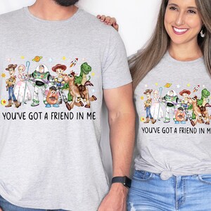 May include: Two light gray t-shirts featuring a colorful graphic of characters from the movie "Toy Story". The text "YOU'VE GOT A FRIEND IN ME" is printed below the graphic. The shirts are worn by two people.