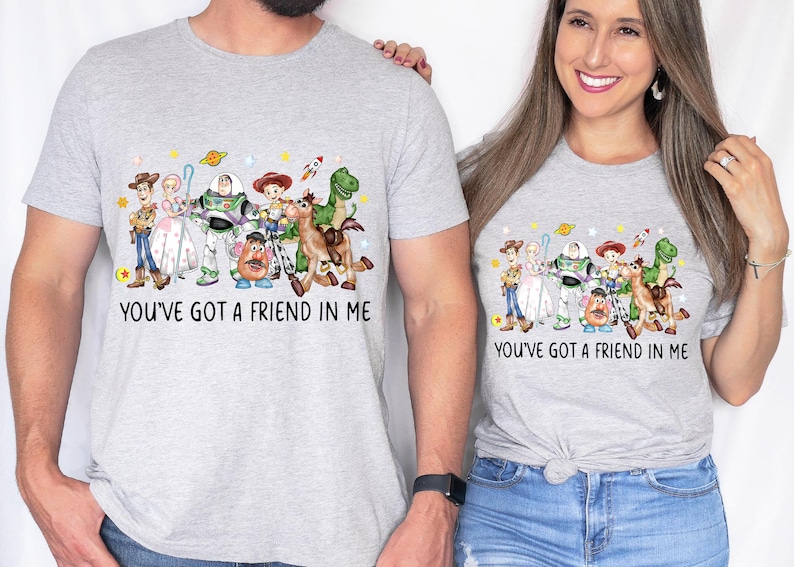May include: Two light gray t-shirts featuring a colorful graphic of characters from the movie "Toy Story". The text "YOU'VE GOT A FRIEND IN ME" is printed below the graphic. The shirts are worn by two people.