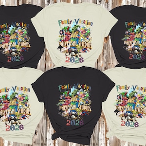 Op de afbeelding: Zes zwarte en cr&egrave;mekleurige t-shirts worden getoond. Elk shirt heeft een kleurrijke afbeelding met de tekst "Family Vacation Universal Studios 2026" en stripfiguren. De shirts zijn gerangschikt op een houten achtergrond.