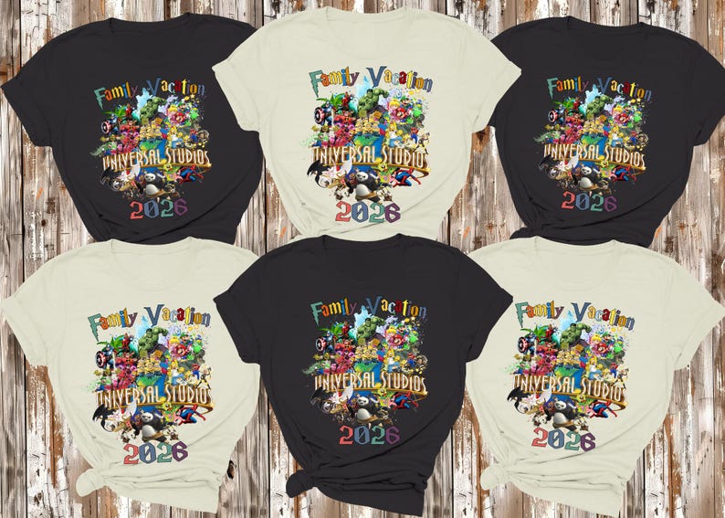 Op de afbeelding: Zes zwarte en cr&egrave;mekleurige t-shirts worden getoond. Elk shirt heeft een kleurrijke afbeelding met de tekst "Family Vacation Universal Studios 2026" en stripfiguren. De shirts zijn gerangschikt op een houten achtergrond.