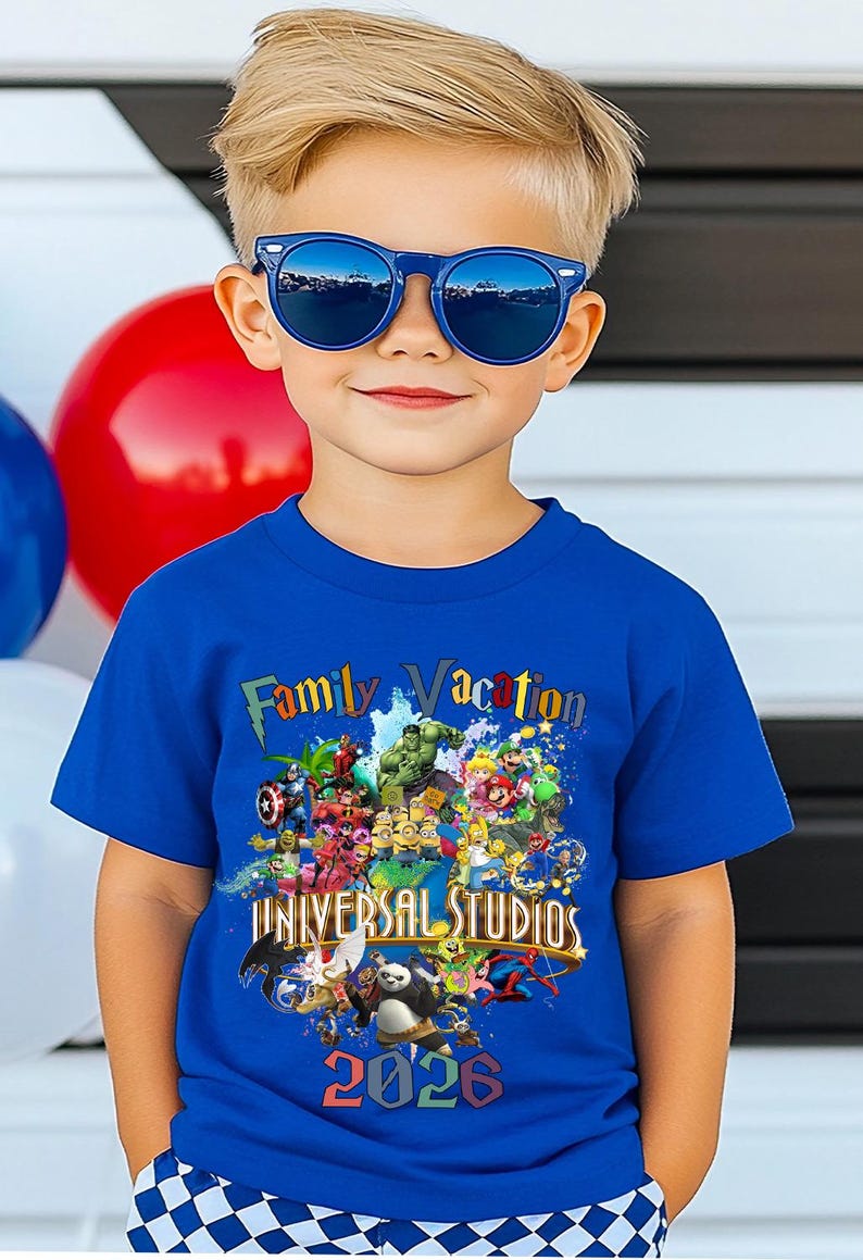Op de afbeelding: Een kind draagt een blauwe zonnebril en een blauw t-shirt. Het shirt heeft een kleurrijke afbeelding met de tekst "Family Vacation" en "Universal Studios 2026", met stripfiguren.