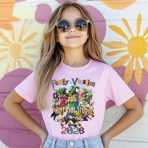 Op de afbeelding: Lichtroze T-shirt met de tekst "Family Vacation" en "Universal Studios 2026", versierd met stripfiguren. Het meisje draagt een zonnebril. De achtergrond is een kleurrijke muurschildering.