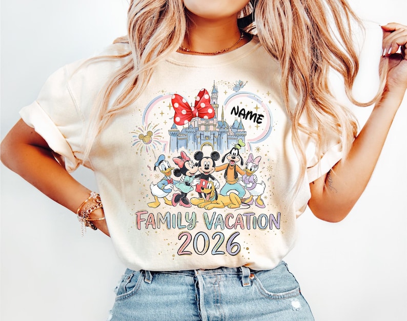 May include: A cream-colored t-shirt featuring a graphic of a castle, a red polka-dot bow, and cartoon characters including Mickey, Minnie, Donald, Daisy, Goofy, and Pluto. The text reads "NAME", "FAMILY VACATION", and "2026" in colorful lettering.