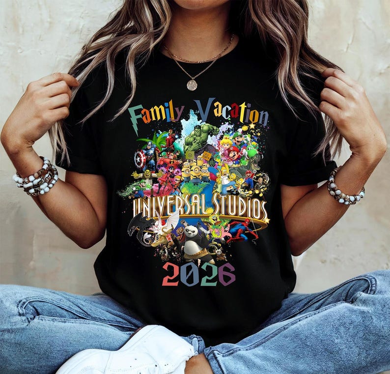 Op de afbeelding: Zwart T-shirt met een kleurrijke afbeelding met de tekst "Family Vacation" en "Universal Studios 2026". De afbeelding bevat verschillende stripfiguren. Het shirt heeft korte mouwen.