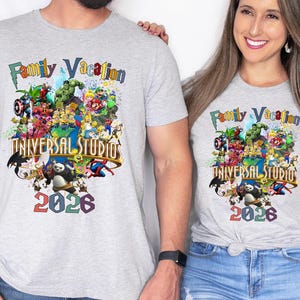 Op de afbeelding: Twee lichtgrijze t-shirts met een kleurrijke afbeelding met verschillende stripfiguren, de tekst "Family Vacation" en "Universal Studios 2026". De shirts hebben korte mouwen.