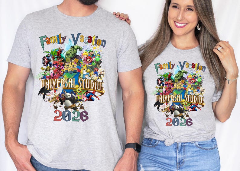 Op de afbeelding: Twee lichtgrijze t-shirts met een kleurrijke afbeelding met verschillende stripfiguren, de tekst "Family Vacation" en "Universal Studios 2026". De shirts hebben korte mouwen.