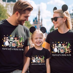 May include: Black t-shirts featuring a colorful graphic of Toy Story characters. The text "YOU'VE GOT A FRIEND IN ME" is printed below the graphic. The shirts are worn by a family, suggesting a theme of family and friendship.