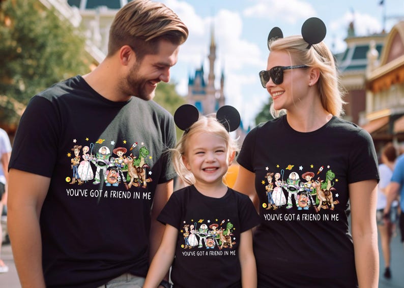 May include: Black t-shirts featuring a colorful graphic of Toy Story characters. The text "YOU'VE GOT A FRIEND IN ME" is printed below the graphic. The shirts are worn by a family, suggesting a theme of family and friendship.
