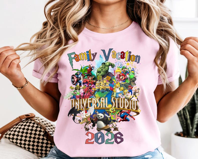 Op de afbeelding: Lichtroze T-shirt met een kleurrijke print. Het ontwerp bevat de woorden "Family Vacation" en "Universal Studios 2026", samen met stripfiguren. Het T-shirt heeft korte mouwen.