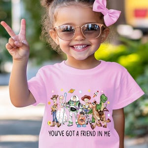 May include: A pink t-shirt featuring a colorful graphic of characters from the animated movie "Toy Story." The shirt has the phrase "YOU'VE GOT A FRIEND IN ME" printed below the characters. The child is wearing sunglasses and a pink bow.