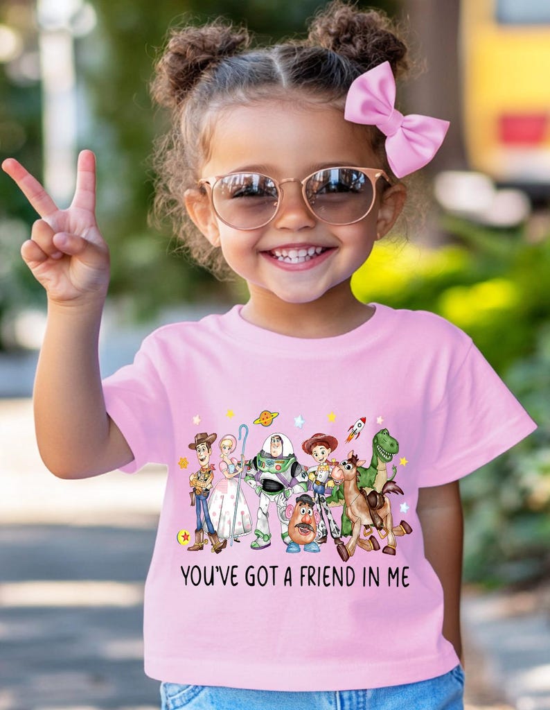 May include: A pink t-shirt featuring a colorful graphic of characters from the animated movie "Toy Story." The shirt has the phrase "YOU'VE GOT A FRIEND IN ME" printed below the characters. The child is wearing sunglasses and a pink bow.