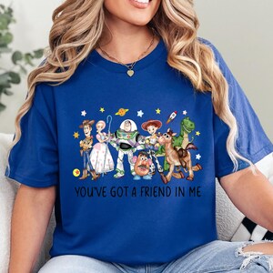 May include: A royal blue t-shirt featuring a colorful graphic of Toy Story characters, including Woody, Buzz Lightyear, and others. The text "YOU'VE GOT A FRIEND IN ME" is printed below the characters. The shirt is a comfortable, casual style.