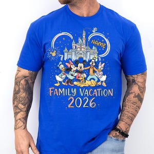 May include: A royal blue short-sleeve t-shirt featuring a colorful graphic of Disney characters including Mickey, Minnie, Donald, Daisy, Goofy, and Pluto in front of a castle. The text reads "NAME", "FAMILY VACATION", and "2026" in a playful font.