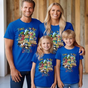 Op de afbeelding: Vier mensen dragen bijpassende blauwe t-shirts met de tekst "Family Vacation UNIVERSAL STUDIOS 2026" en een kleurrijke afbeelding. De shirts hebben korte mouwen en de familie lacht.
