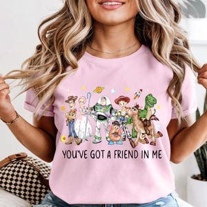 May include: A light pink t-shirt featuring a colorful graphic of characters from the animated movie Toy Story. The graphic includes Woody, Buzz Lightyear, and other characters, with the text "YOU'VE GOT A FRIEND IN ME" below.