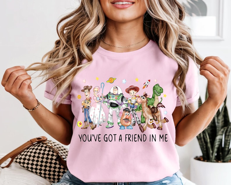 May include: A light pink t-shirt featuring a colorful graphic of characters from the animated movie Toy Story. The graphic includes Woody, Buzz Lightyear, and other characters, with the text "YOU'VE GOT A FRIEND IN ME" below.