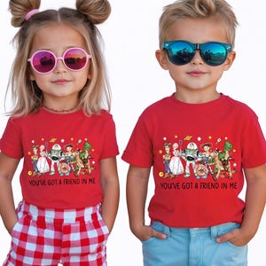 May include: Two children wearing red t-shirts with a colorful graphic of animated characters and the text "YOU'VE GOT A FRIEND IN ME". The girl wears pink sunglasses and the boy wears blue sunglasses.