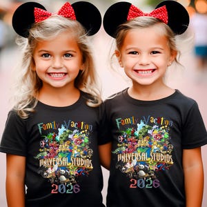 Op de afbeelding: Twee jonge meisjes dragen zwarte t-shirts met een kleurrijk grafisch ontwerp en de tekst "Family Vacation UNIVERSAL STUDIOS 2026". Ze dragen ook zwarte Mickey Mouse oren met rode stippen strikken.