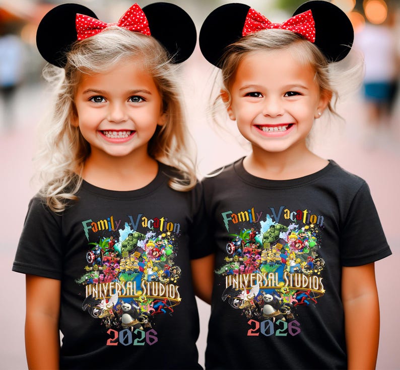 Op de afbeelding: Twee jonge meisjes dragen zwarte t-shirts met een kleurrijk grafisch ontwerp en de tekst "Family Vacation UNIVERSAL STUDIOS 2026". Ze dragen ook zwarte Mickey Mouse oren met rode stippen strikken.