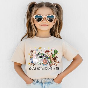 May include: A beige t-shirt featuring a colorful graphic of characters from the animated film "Toy Story." The graphic includes Woody, Buzz Lightyear, and other characters. The text "YOU'VE GOT A FRIEND IN ME" is printed below the characters.