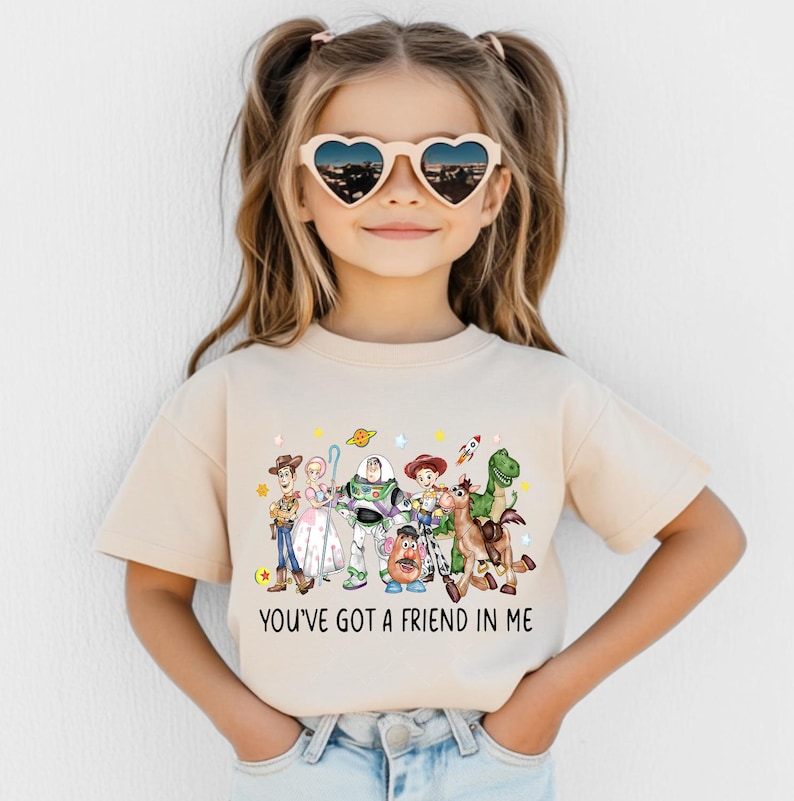 May include: A beige t-shirt featuring a colorful graphic of characters from the animated film "Toy Story." The graphic includes Woody, Buzz Lightyear, and other characters. The text "YOU'VE GOT A FRIEND IN ME" is printed below the characters.