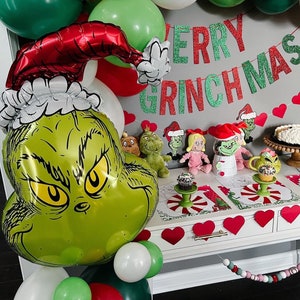 Pu&ograve; includere: Un palloncino verde Grinch con un cappello rosso e un bordo di pelliccia bianca. Il palloncino &egrave; circondato da palloncini verdi, rossi e bianchi. Uno striscione di glitter verde e rosso recita "Merry Grinchmas".
