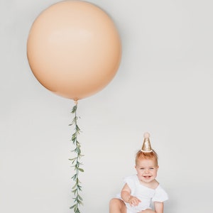 May include: A baby wearing a white dress and a gold glitter party hat sits on a white background. A large peach balloon with a green garland hangs above the baby.