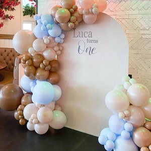 Op de afbeelding: Een witte boog achtergrond versierd met een ballon slinger in tinten bruin, beige en blauw. De slinger is gemaakt van verschillende maten ballonnen. De achtergrond heeft de tekst "Luca turns One" erop.