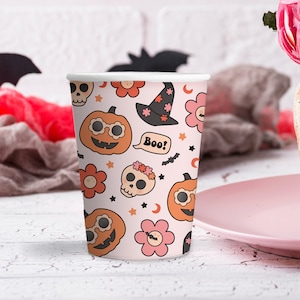 Może przedstawiać: Papierowy kubek z wzorem Halloween, przedstawiający dynie, kapelusze czarownic, kwiaty, czaszki i napis "Boo!" na jasnor&oacute;żowym tle. G&oacute;ra i d&oacute;ł kubka są białe.