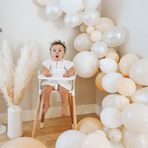 Może przedstawiać: Mała dziewczynka w białej sukience i opasce z kwiatami siedzi na białym wysokim krzesełku. Krzesło ma drewniane nogi. Dziewczynka jest otoczona girlandą kremowych i białych balon&oacute;w.