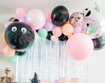May include: A Halloween party table setting with a black and pink witch hat, black and silver disco balls, and a white pumpkin. The table is decorated with a black tablecloth with white text that says "Dead is just the start of the fun". There is a silver garland of balloons above the table, including black, pink, and green balloons.