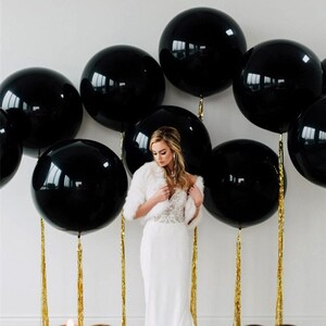 Puede incluir: Un vestido blanco con un corpi&ntilde;o de encaje y una estola de piel blanca delante de una pared decorada con globos negros. Los globos est&aacute;n atados con serpentinas doradas.