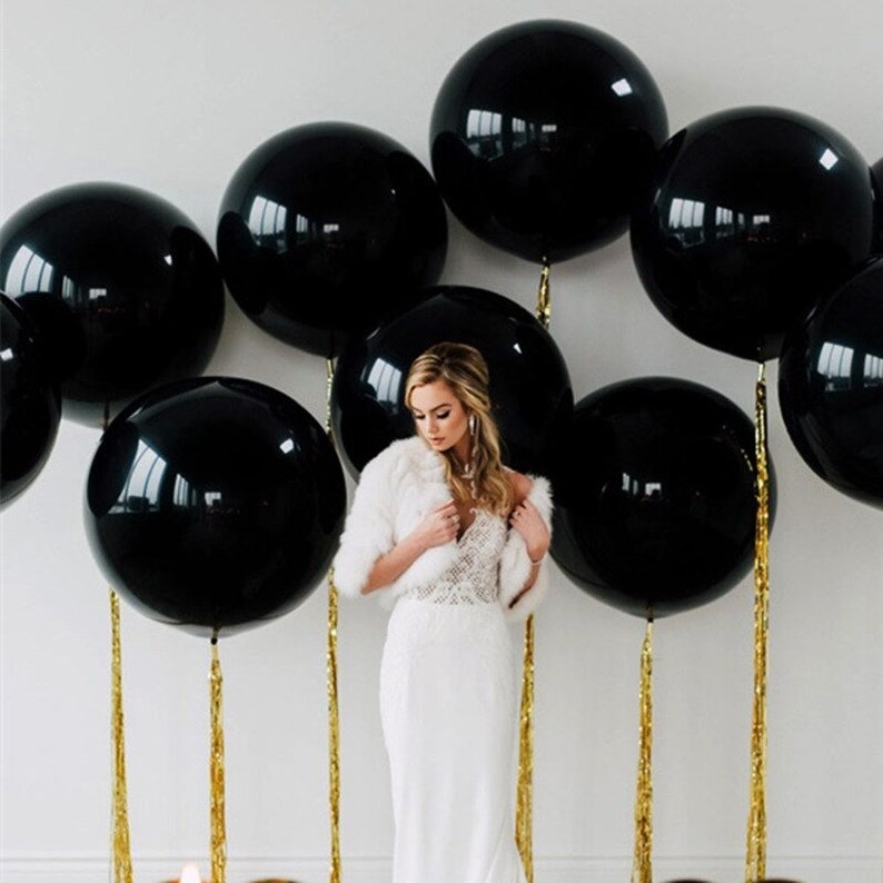 Puede incluir: Un vestido blanco con un corpi&ntilde;o de encaje y una estola de piel blanca delante de una pared decorada con globos negros. Los globos est&aacute;n atados con serpentinas doradas.