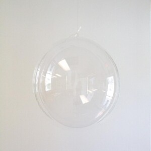 Op de afbeelding: Een doorzichtige plastic bubbelballon die aan een touwtje hangt. De ballon is opgeblazen en lijkt rond te zijn.