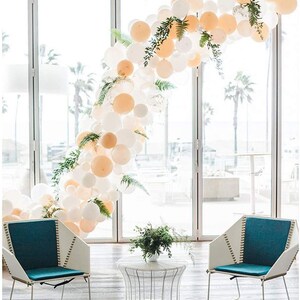 Puede incluir: Un arco de globos blanco y melocot&oacute;n con vegetaci&oacute;n, dos sillas de color azul verdoso y blanco y una mesa de metal blanco con un jarr&oacute;n de vegetaci&oacute;n en la parte superior. El arco de globos est&aacute; delante de una ventana grande con vistas a palmeras.