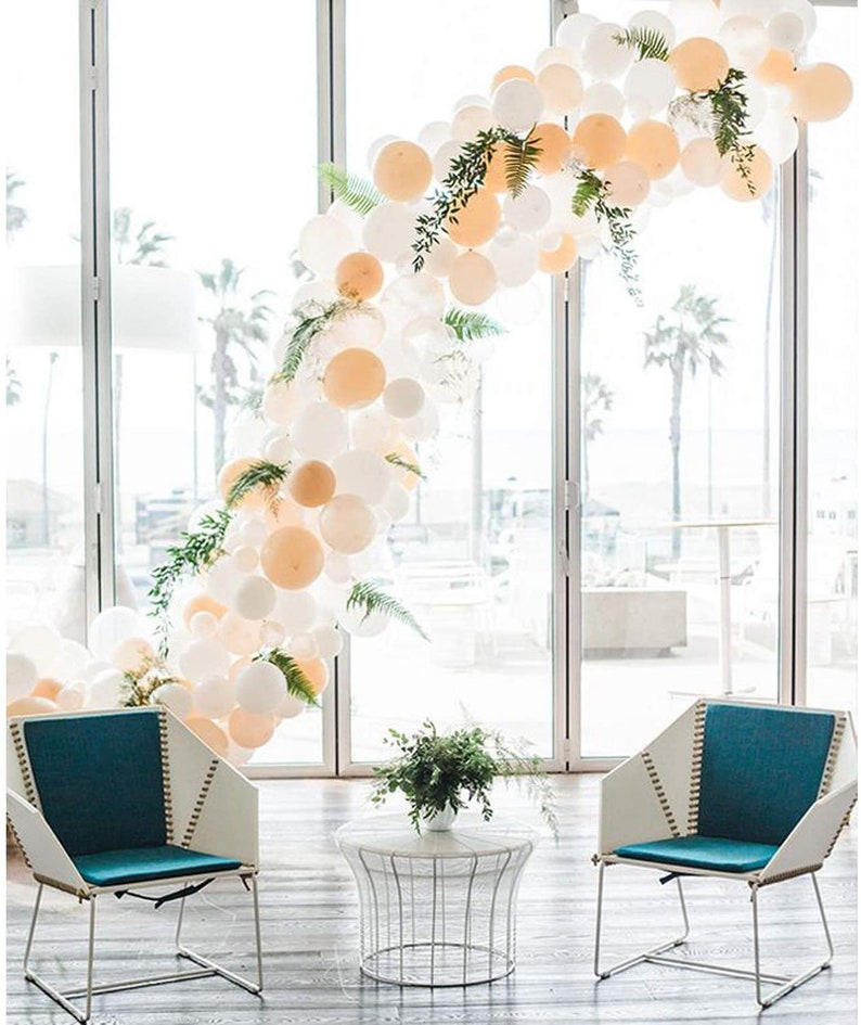 Puede incluir: Un arco de globos blanco y melocot&oacute;n con vegetaci&oacute;n, dos sillas de color azul verdoso y blanco y una mesa de metal blanco con un jarr&oacute;n de vegetaci&oacute;n en la parte superior. El arco de globos est&aacute; delante de una ventana grande con vistas a palmeras.